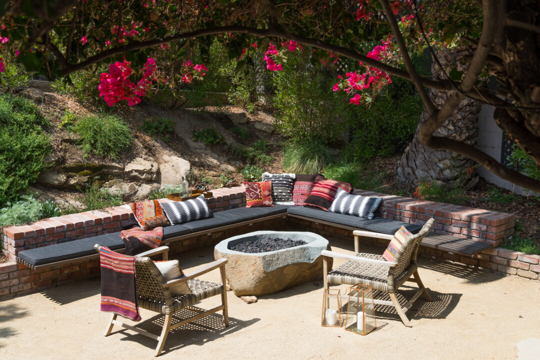 A stone fire pit beside an L shaped outdoor couch, beside a hillside